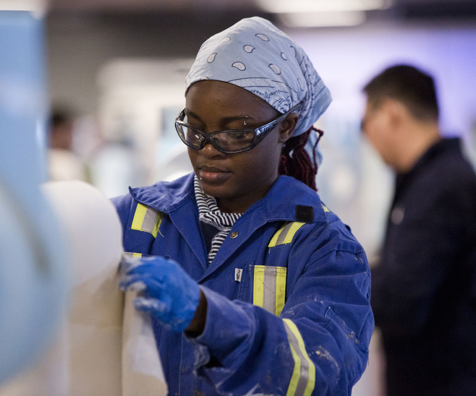A woman in blue coveralls and safety glasses is engaged in a task, showcasing her focus and attention in a collaborative environment.