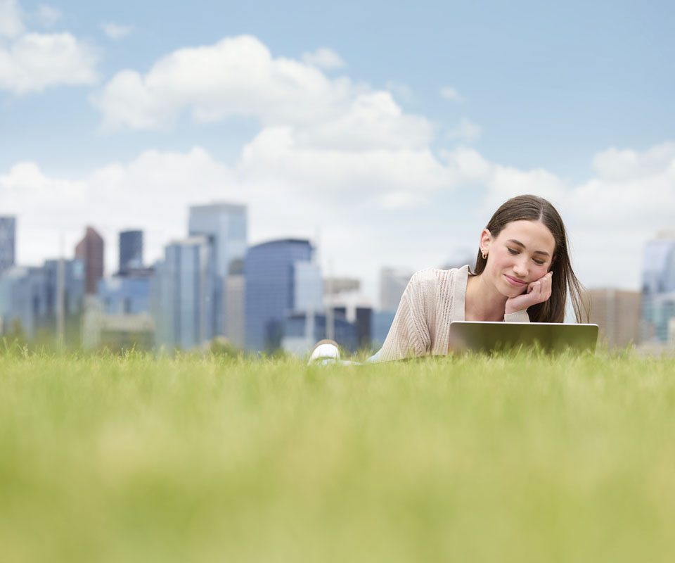 A young woman lies on the grass with a laptop, smiling and enjoying her work against a city skyline backdrop.