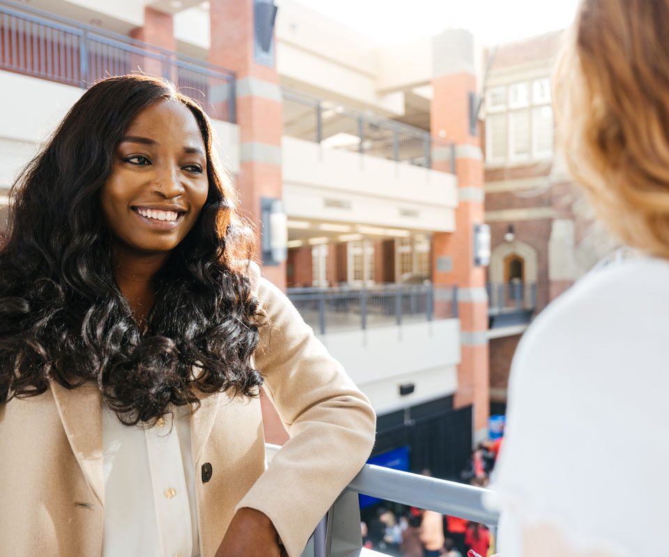A woman with long, wavy hair smiles while speaking to another person in a sunny indoor setting.