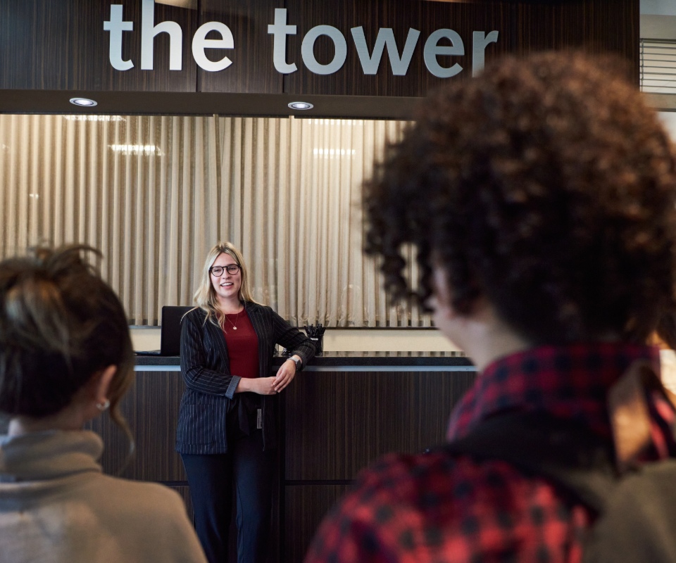 A smiling receptionist stands at the front desk welcoming two visitors.