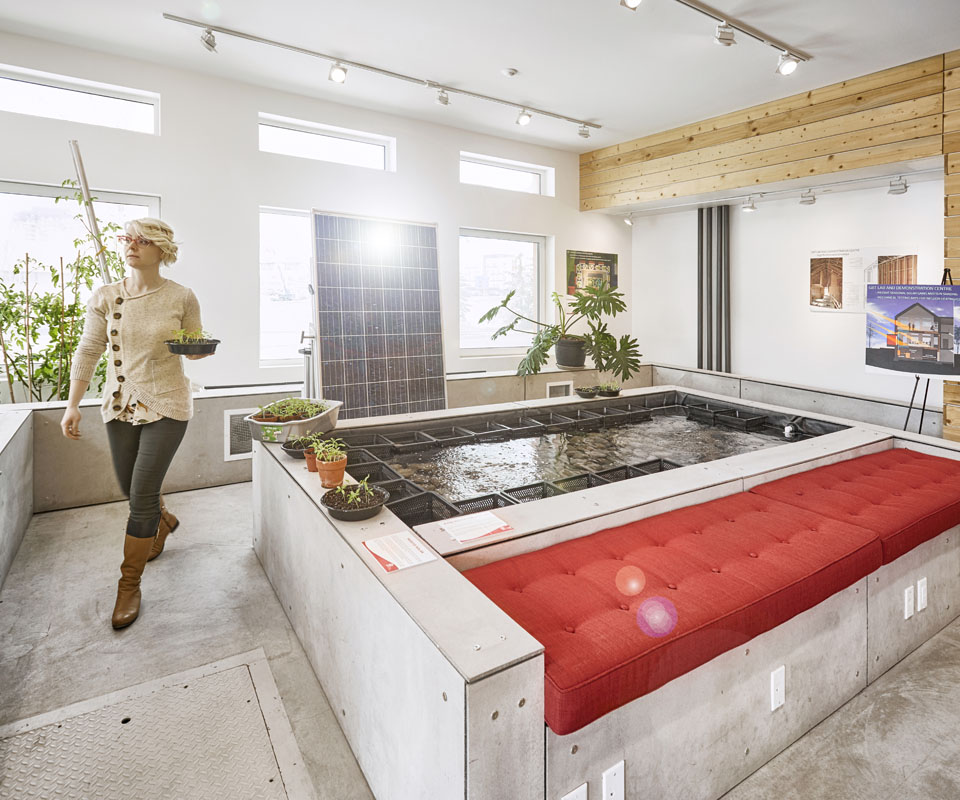 A modern indoor aquaponics system with a woman carrying plants, featuring a sunlit room with large windows and a concrete bench.