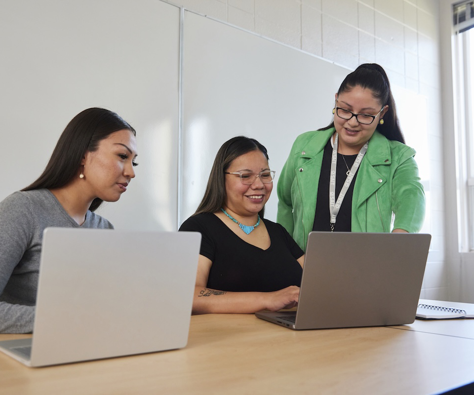 Three Indigenous women collaborating on laptops in a classroom setting, with one woman guiding the others.