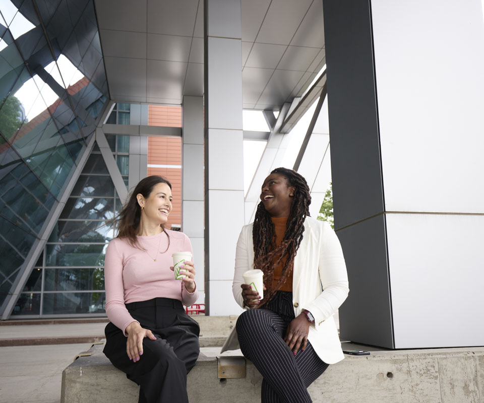 Two women sit outdoors enjoying coffee while engaged in conversation, with the modern Aldred Centre in the background.