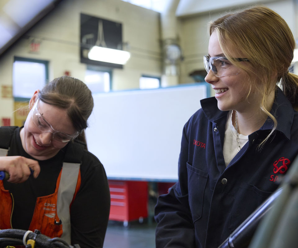 Two women in safety goggles working together in a workshop, smiling and engaging in hands-on mechanical tasks.