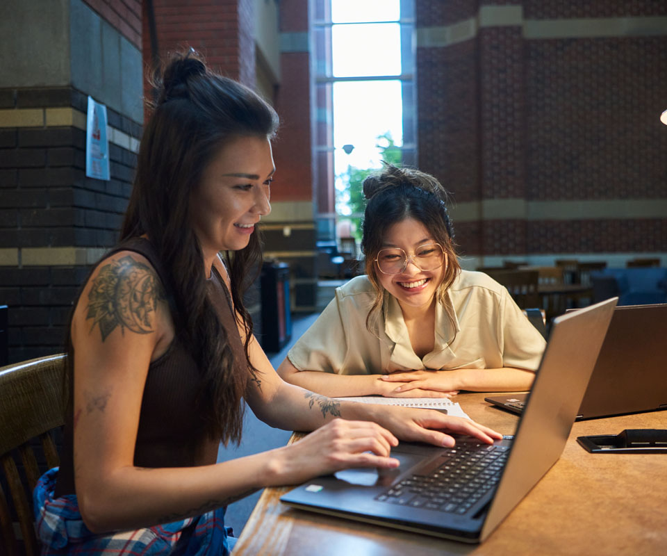 Two young women collaborate and smile while working on a laptop in a bright study environment. Their engaged expressions convey a sense of teamwork and joy in learning.