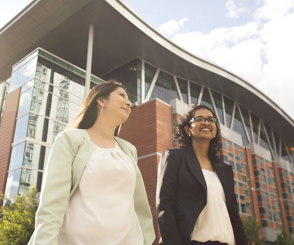 Two professional women walking together outside the modern Aldred Centre building, showcasing a collaborative and positive atmosphere.