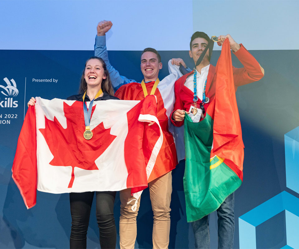 Three smiling WorldSkills competitors celebrating on stage, holding flags of Canada and other countries, showcasing their achievement during an awards ceremony.