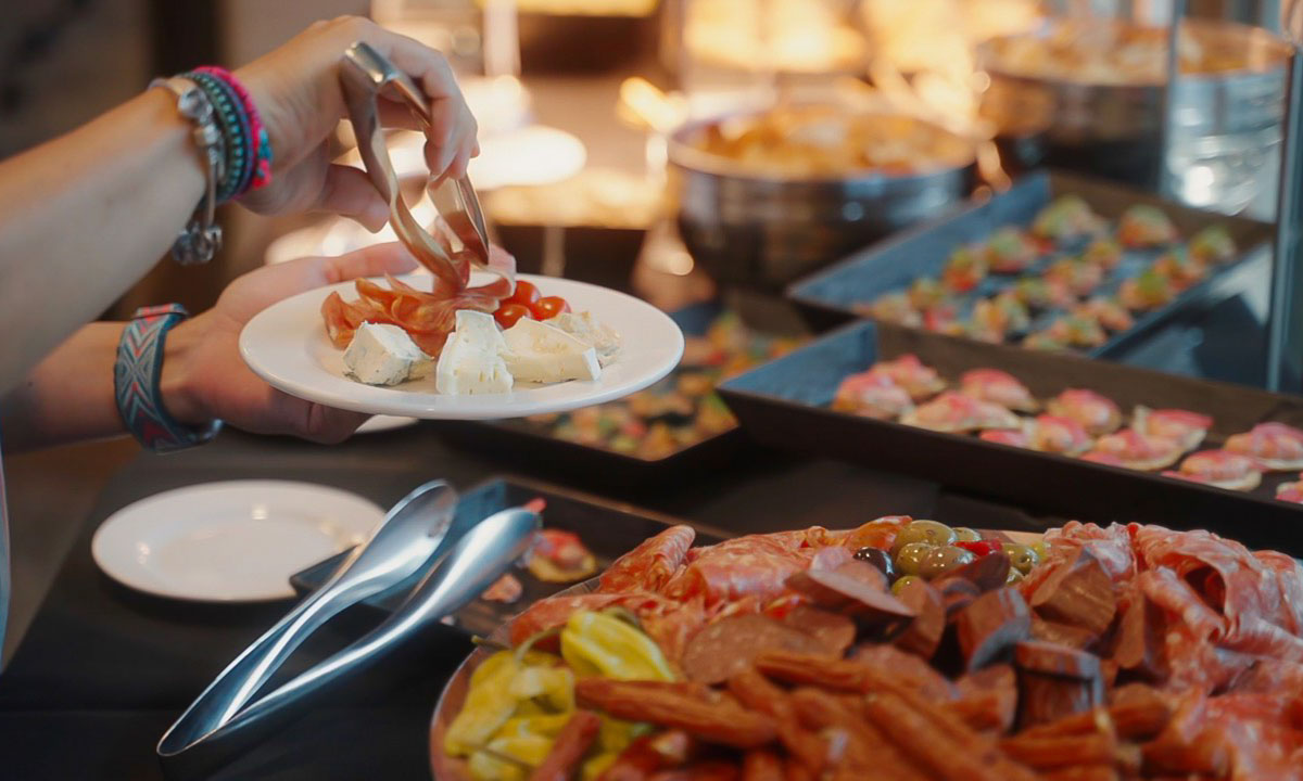 A person serving sliced meats and cheese from a buffet spread.