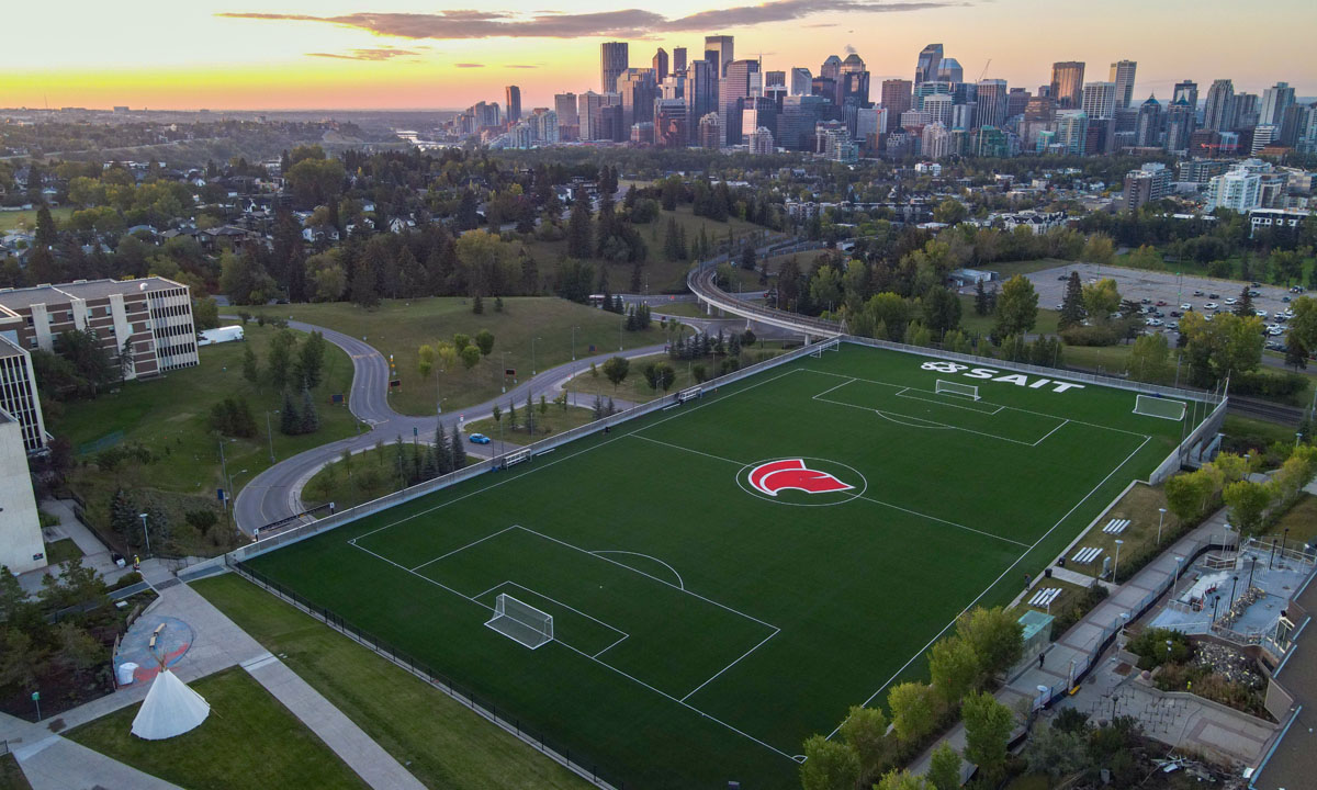 Aerial view of the Cohos Commons soccer field featuring a large SAIT logo, surrounded by urban cityscape and green parks in the background.
