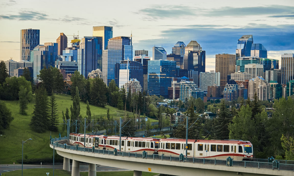 A c-train travels along a curved bridge with the Calgary skyline in the background, showcasing a mix of modern skyscrapers against a green landscape.
