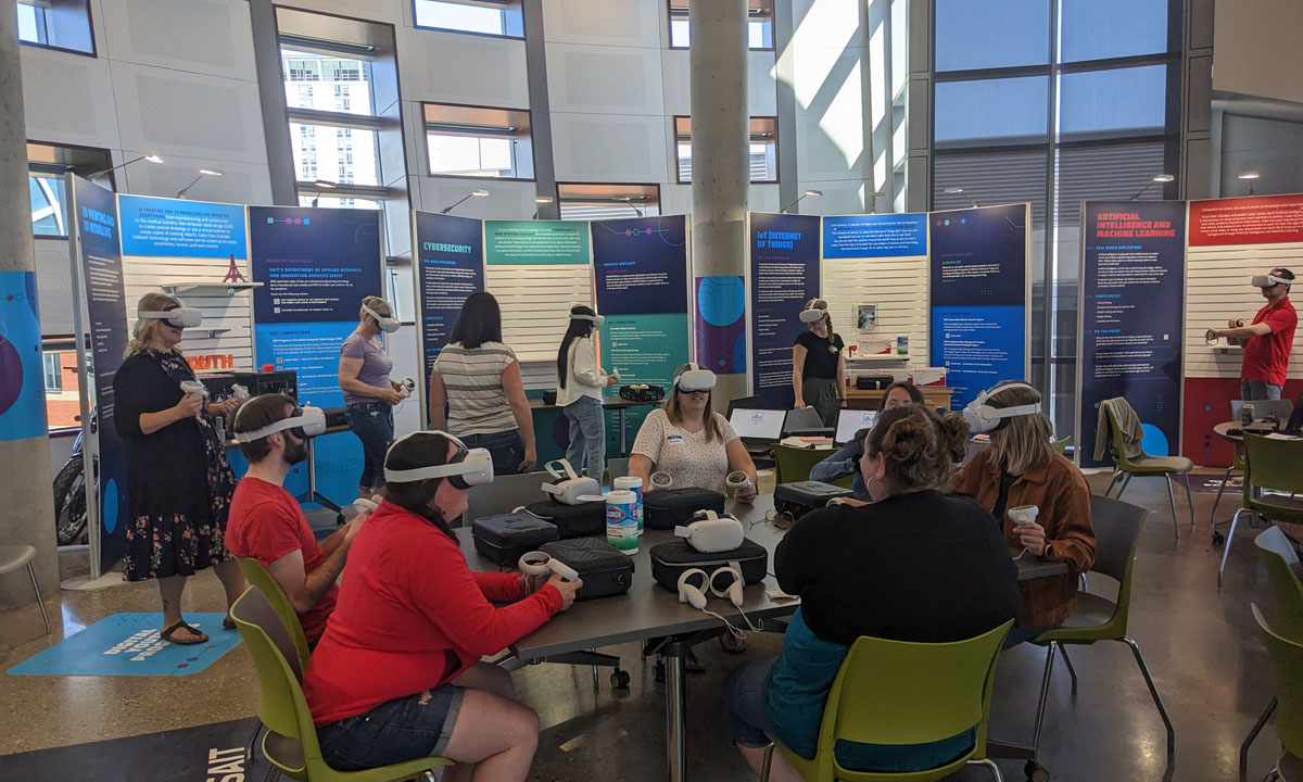 A group of high school students in a modern classroom setting use virtual reality headsets and controllers, participating in an interactive digital learning activity surrounded by colourful informational displays.