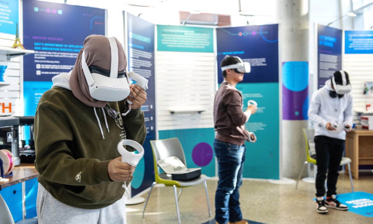 A group of students engaging with virtual reality headsets in an interactive exhibit space. Informational panels are displayed in the background.