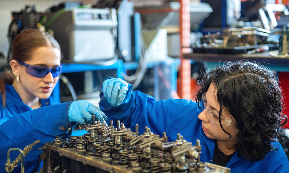 Two high-school dual-credit students working together on a car engine in an automotive workshop.