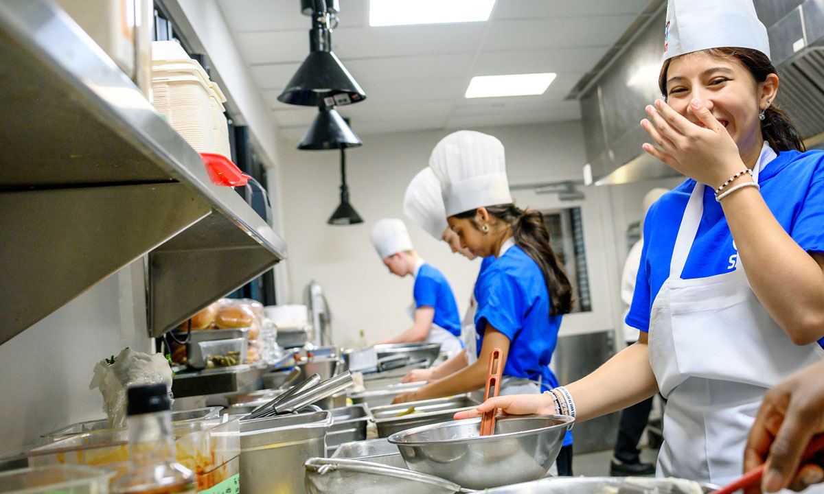 Young adults in a bustling kitchen environment prepare food, showcasing teamwork and culinary skills. One student joyfully laughs while stirring a bowl, highlighting a positive and energetic atmosphere.