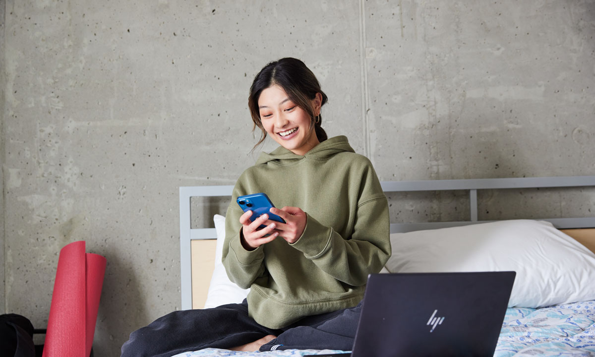 A smiling young woman sits on a bed, using a smartphone, with a laptop nearby in a modern dorm room.