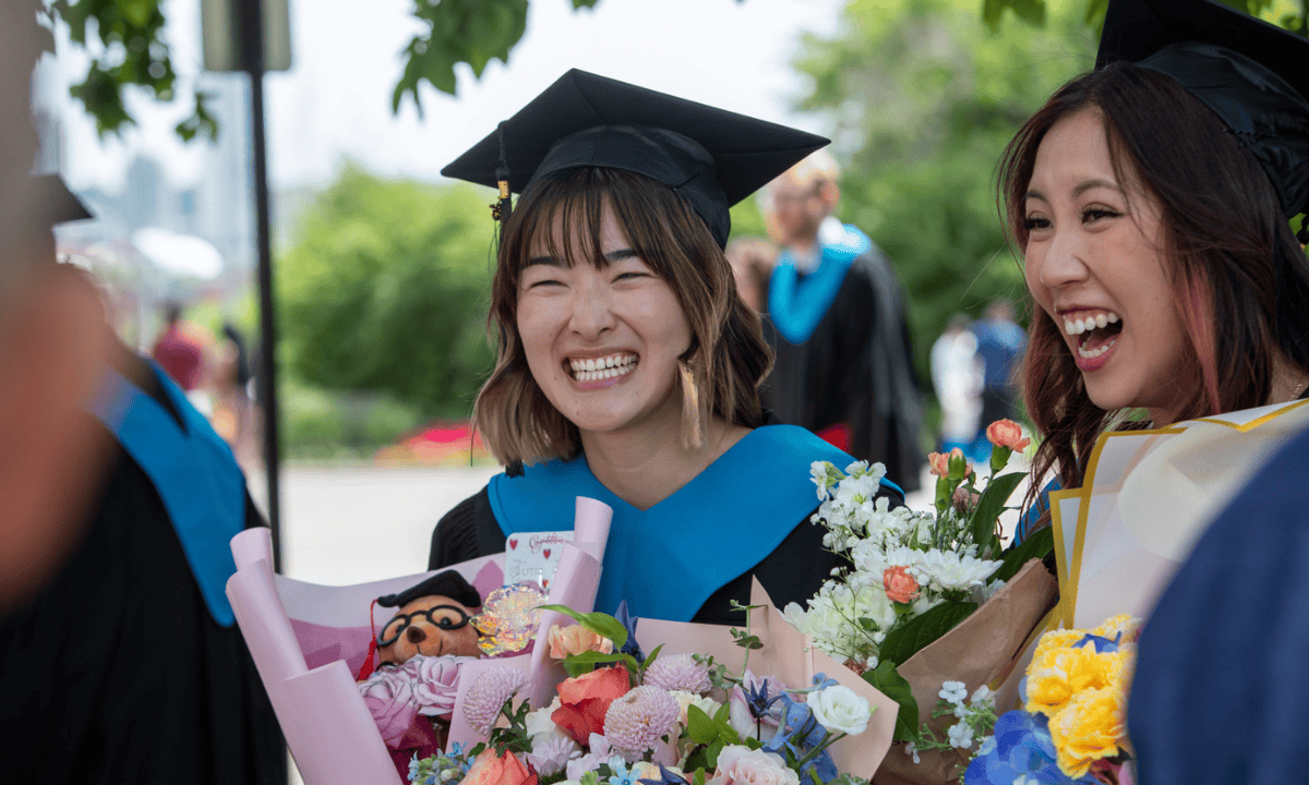 Two smiling graduates in caps and gowns celebrate with floral bouquets.