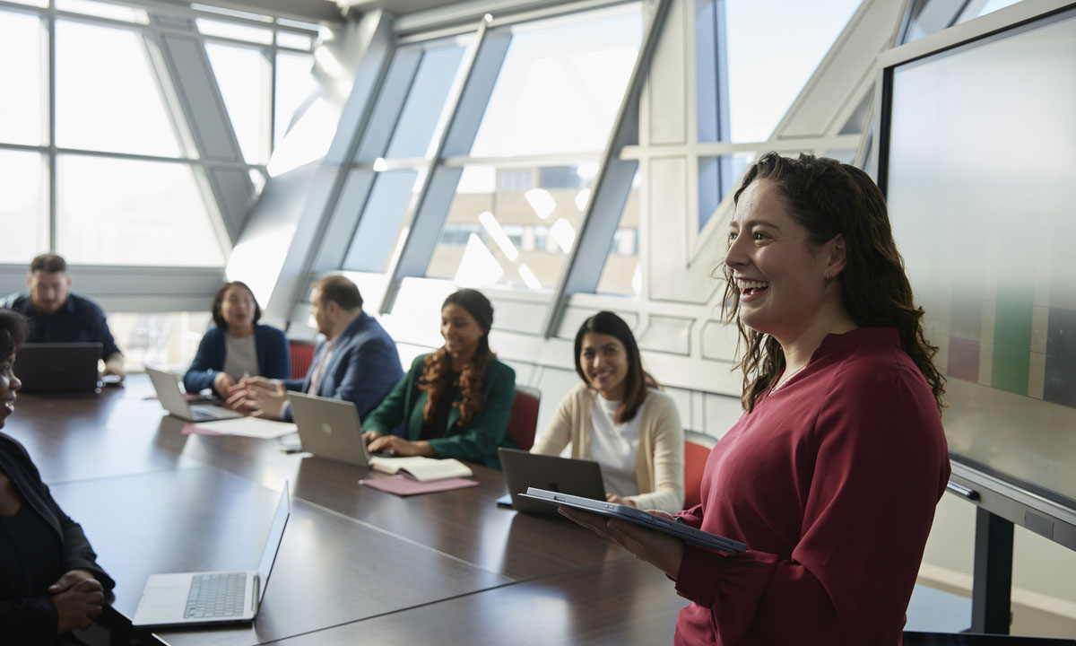 A businesswoman presenting in a modern conference room while colleagues engage around a table with laptops.