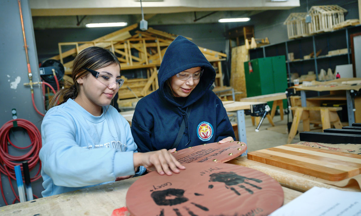 Two Indigenous students wearing safety glasses work together in a woodworking shop, decorating a wooden project with painted handprints and text, surrounded by tools and materials.