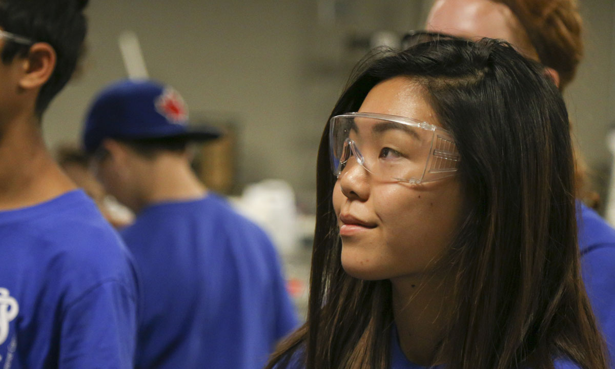 A student wearing safety goggles listens attentively in a classroom or lab setting, with other students in blue shirts working in the background.