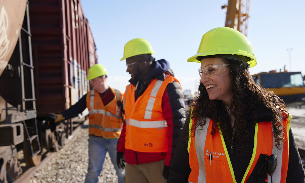 A diverse group of railway workers wearing safety vests and helmets, smiling and engaging at the rail yard at Point Trotter campus.