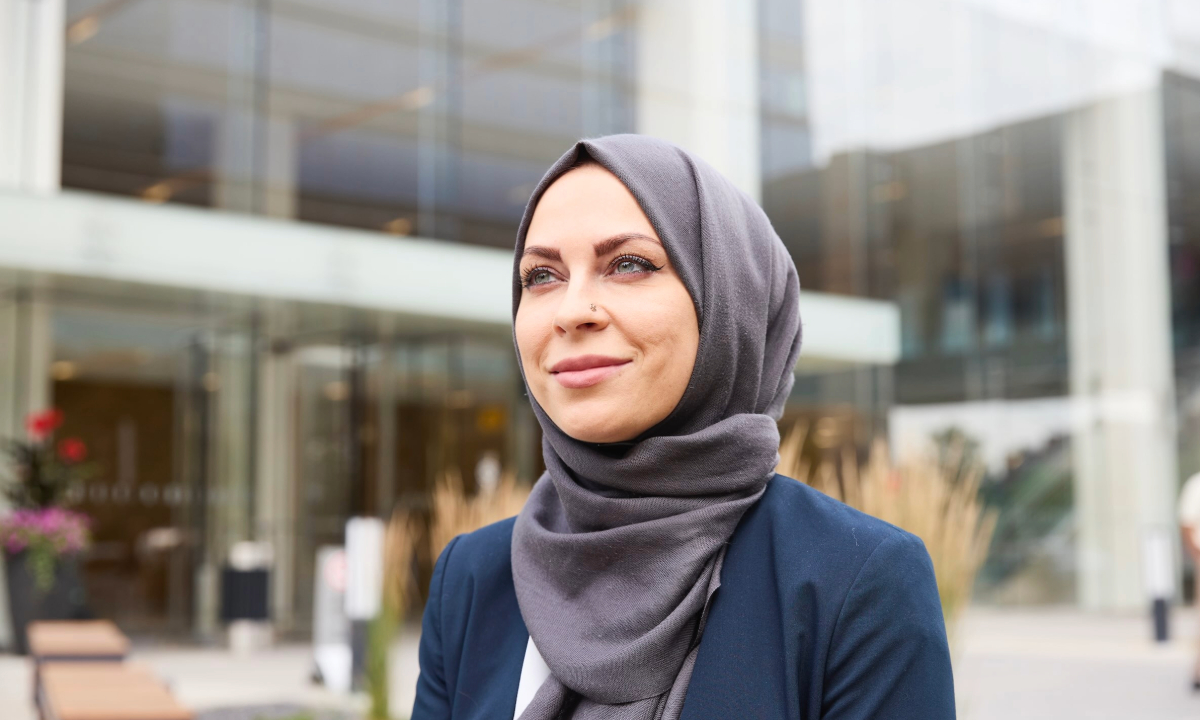 A confident woman wearing a hijab and business attire stands outside a modern building, looking thoughtfully into the distance.