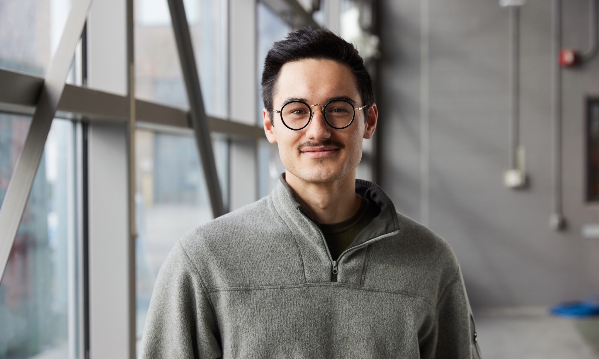 A smiling man with glasses and a mustache is standing indoors, dressed in a gray sweater against a backdrop of large windows.