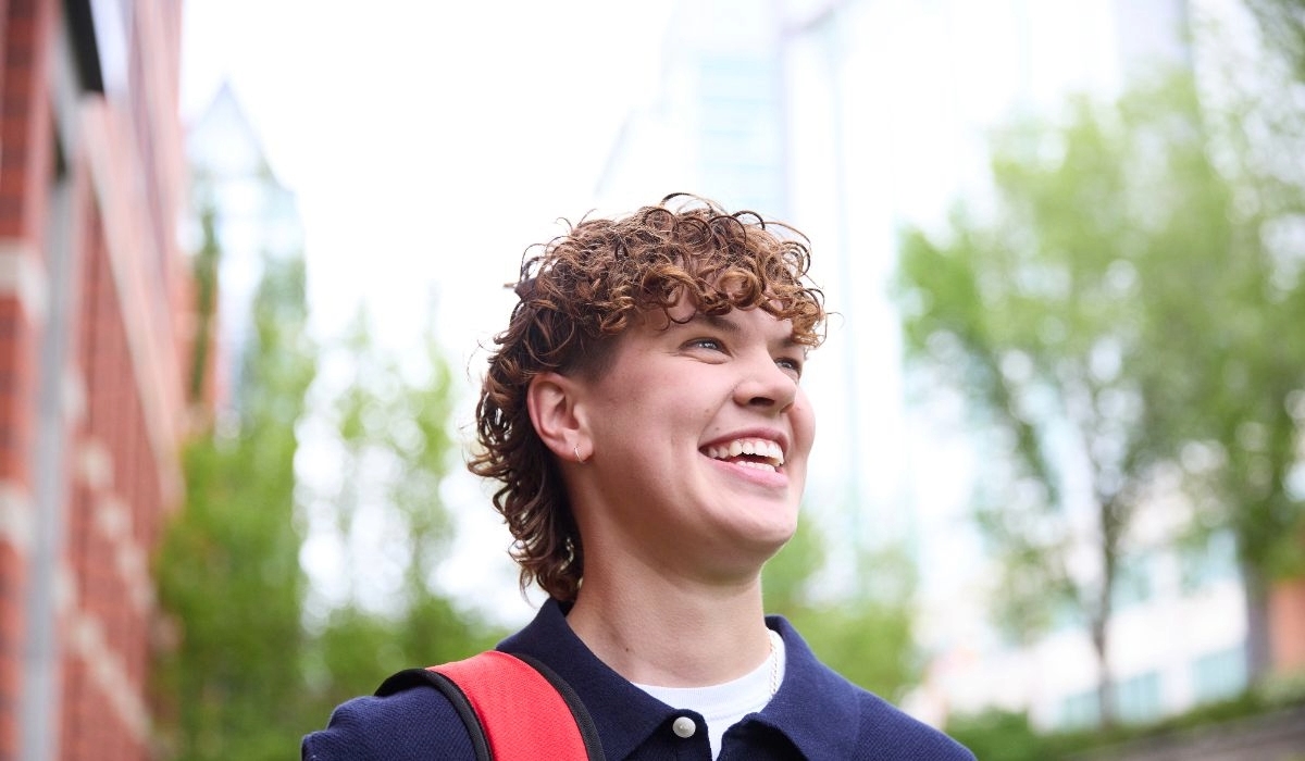 A smiling student with curly hair stands outdoors, wearing a dark shirt and carrying a red backpack.