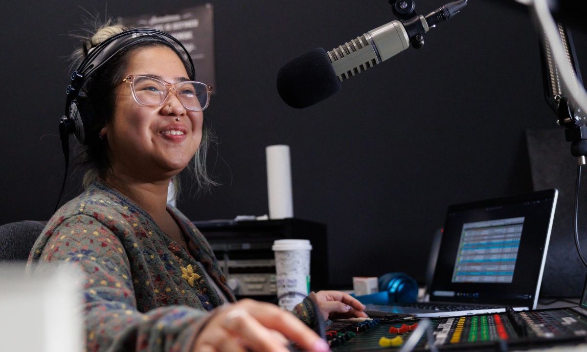 A woman with glasses is smiling while operating a sound mixing console in a radio studio.