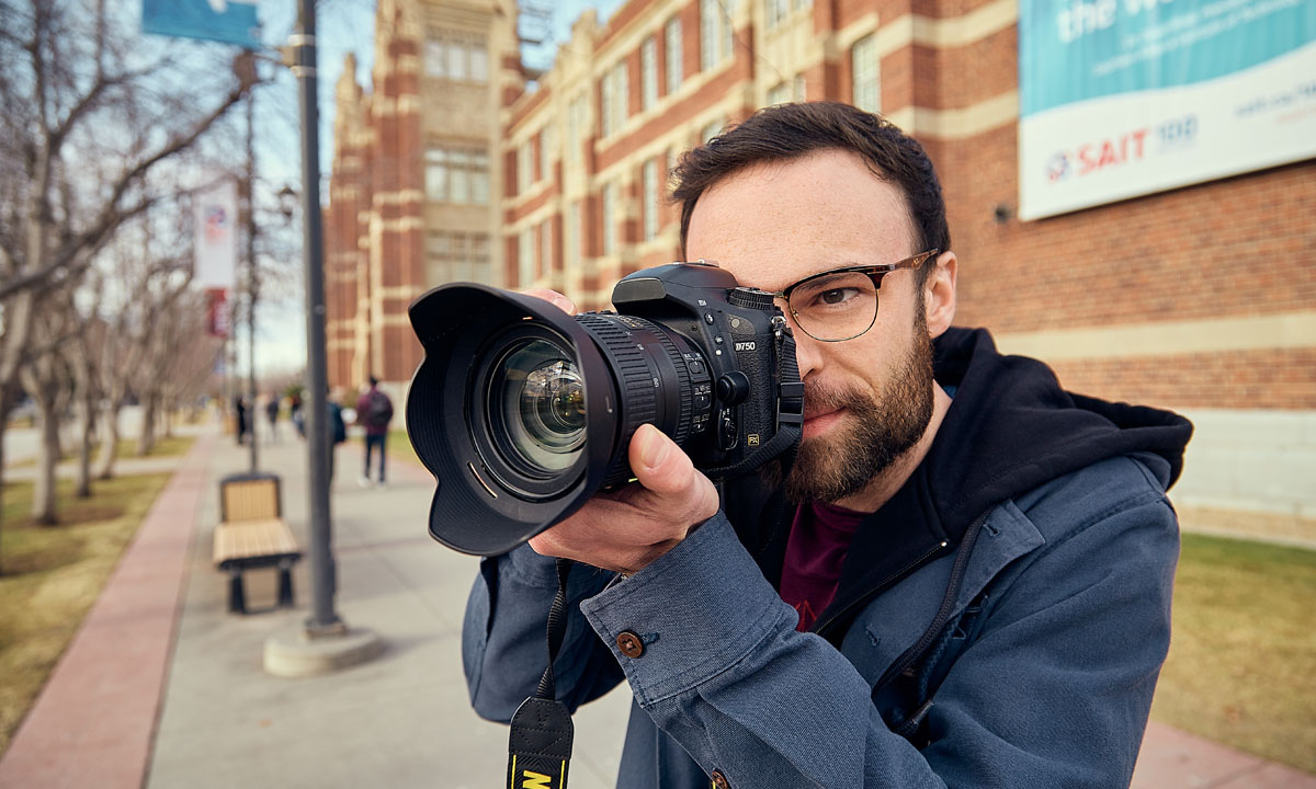 A man with glasses holding a DSLR camera, preparing to take a photo on SAIT campus.