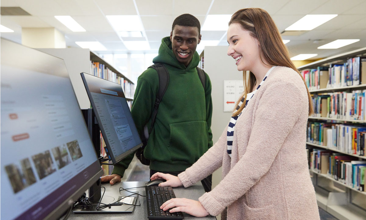 Two students use desktop computers together in a library, smiling as one types on the keyboard.