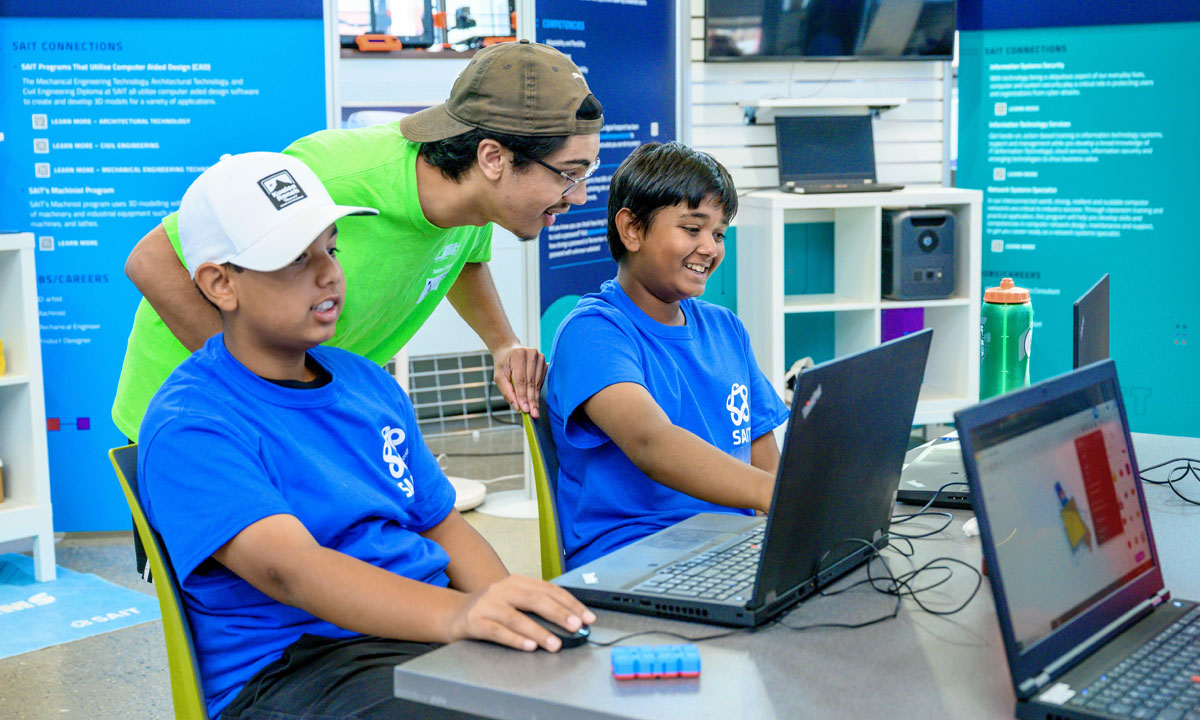 Two summer campers in blue shirts are engaged with laptops, while a camp leader in a green shirt enthusiastically helps them with their project.