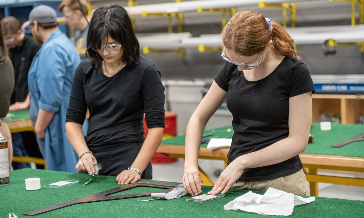 Two students in safety goggles engaged in hands-on work in a workshop, focused on crafting or assembling materials at a workspace.