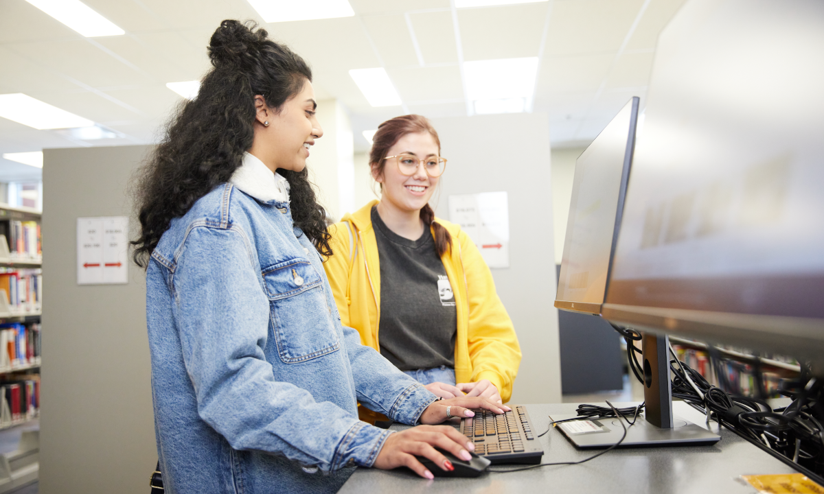 Two young women collaborating at a computer in a library setting. One woman is typing on the keyboard while the other smiles and watches.