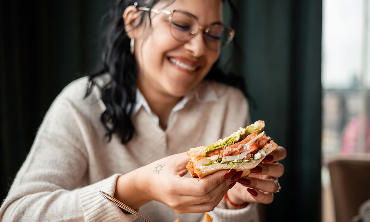 A woman smiling while enjoying a delicious sandwich, surrounded by a cozy dining atmosphere.