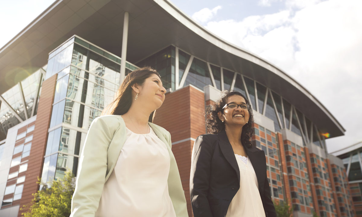Two professional women walking together outside the Aldred Centre, smiling and engaging in conversation.