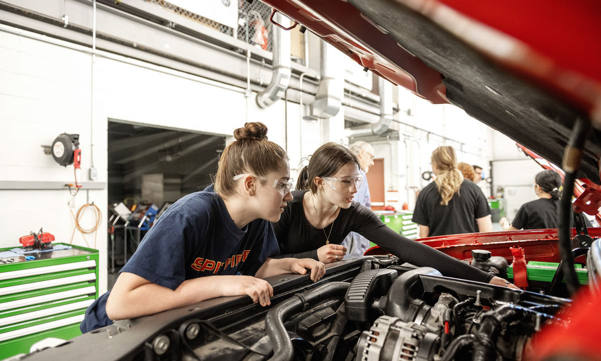 Two young women wearing safety goggles closely examine the engine of a car during a hands-on automotive workshop.