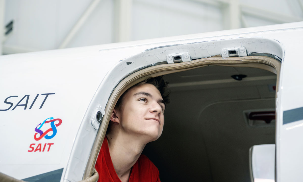 A young man peeks out from the cockpit of an aircraft, showcasing excitement and curiosity. The SAIT logo is visible on the side of the aircraft.