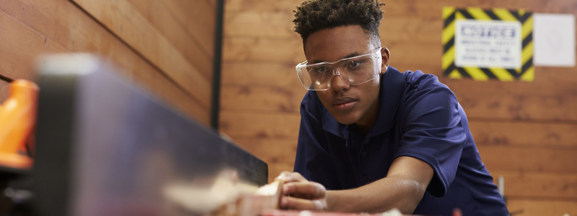 A focused apprentice wearing safety glasses leans over a workbench in a workshop setting.