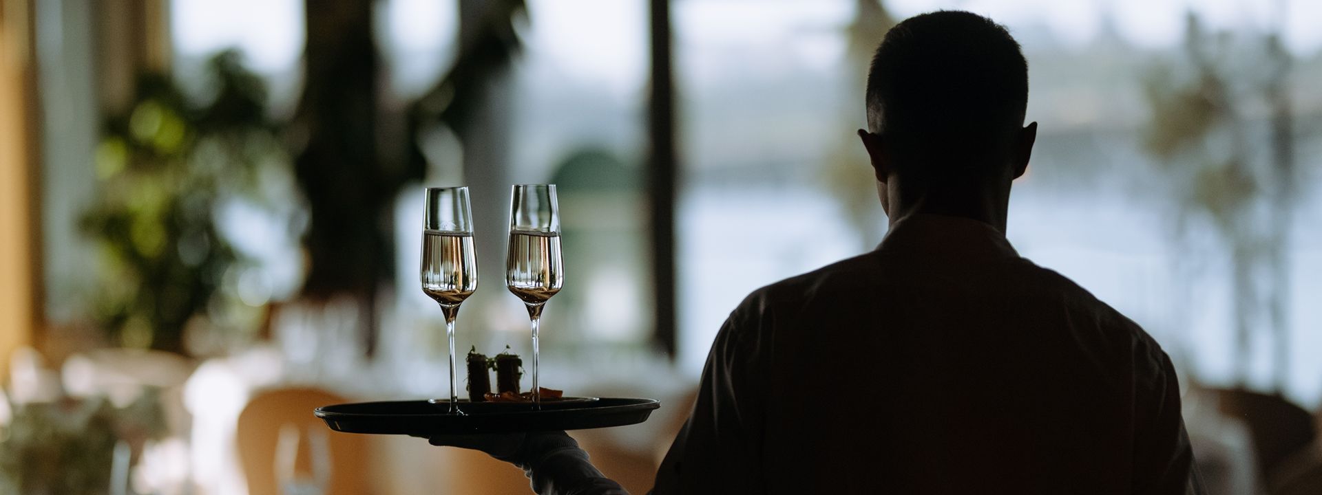 Server carrying two glasses of champagne on a tray in a softly lit restaurant.