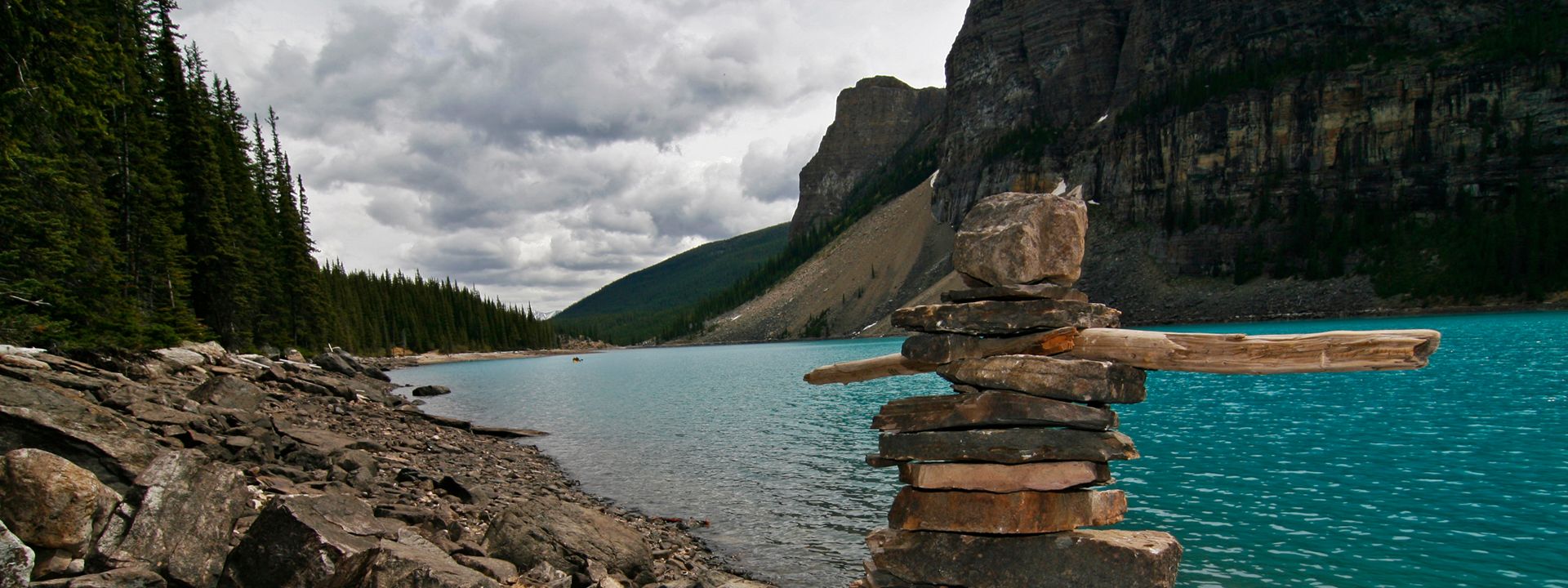 Stacked rocks on a lakeshore with turquoise water, pine trees, and mountains under a cloudy sky.