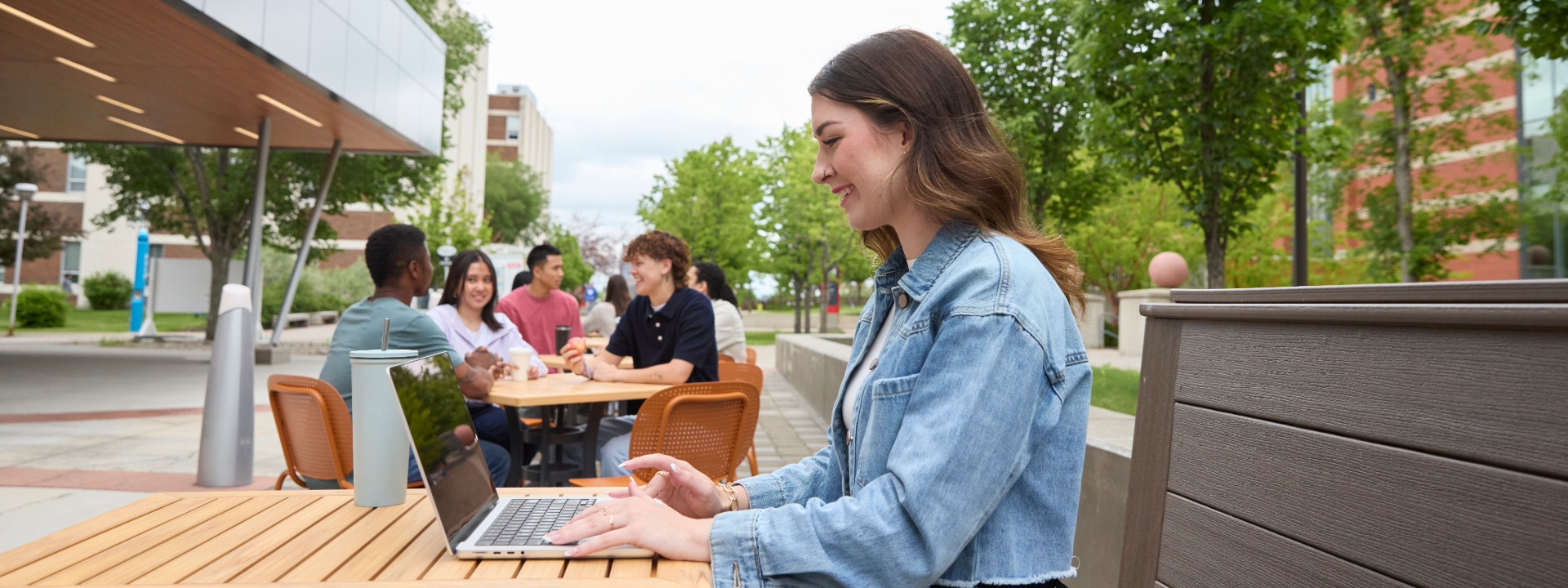 A student works on a laptop outdoors at a campus seating area, with other students socializing in the background.