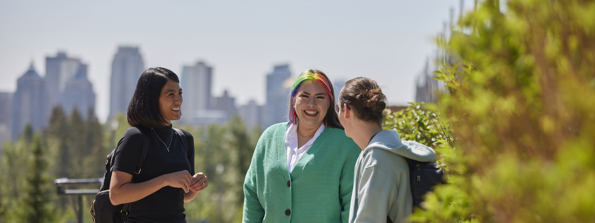Three diverse students are smiling and interacting outdoors, with a city skyline in the background.