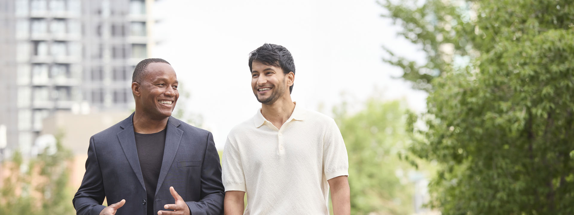 Two men smiling and walking together in an outdoor setting with greenery and modern buildings in the background.