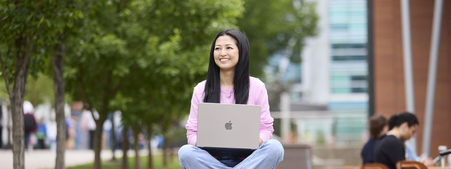 A smiling woman sits outdoors on a grassy area, working on a laptop in her lap. She is surrounded by trees and people in a lively setting.