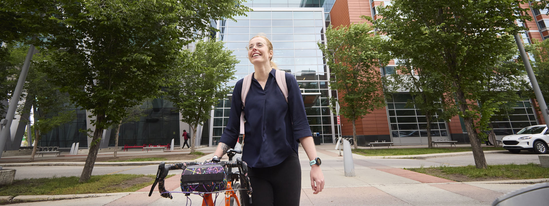 A woman smiling while walking her bicycle outside the Aldred Centre surrounded by trees.