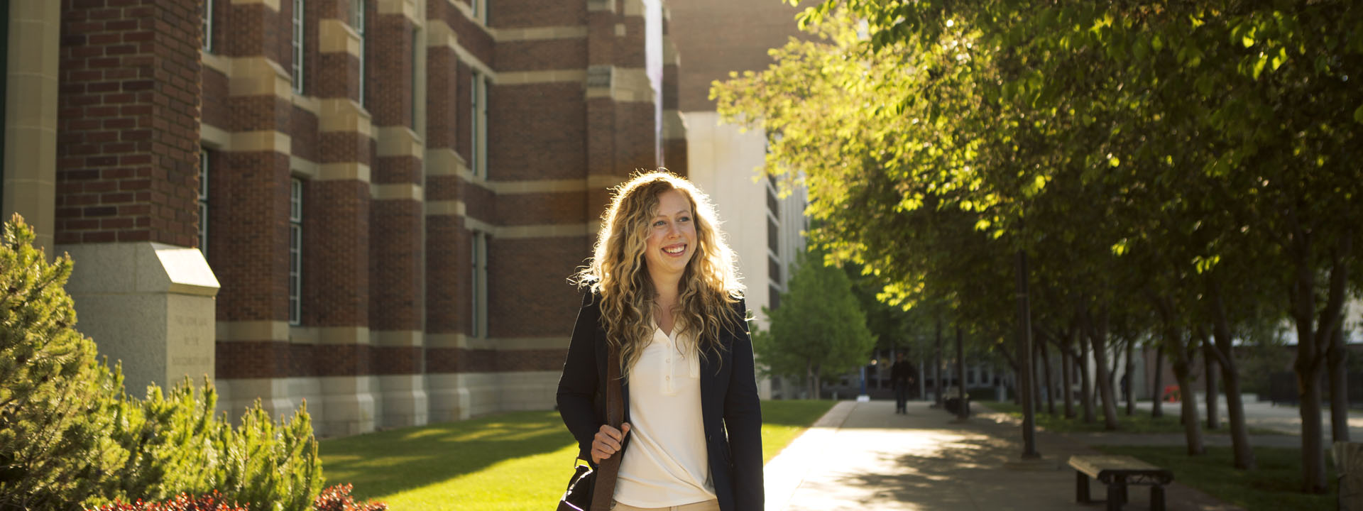 A smiling woman with curly hair walks along a tree-lined path, dressed in business attire, in front of Heritage Hall.