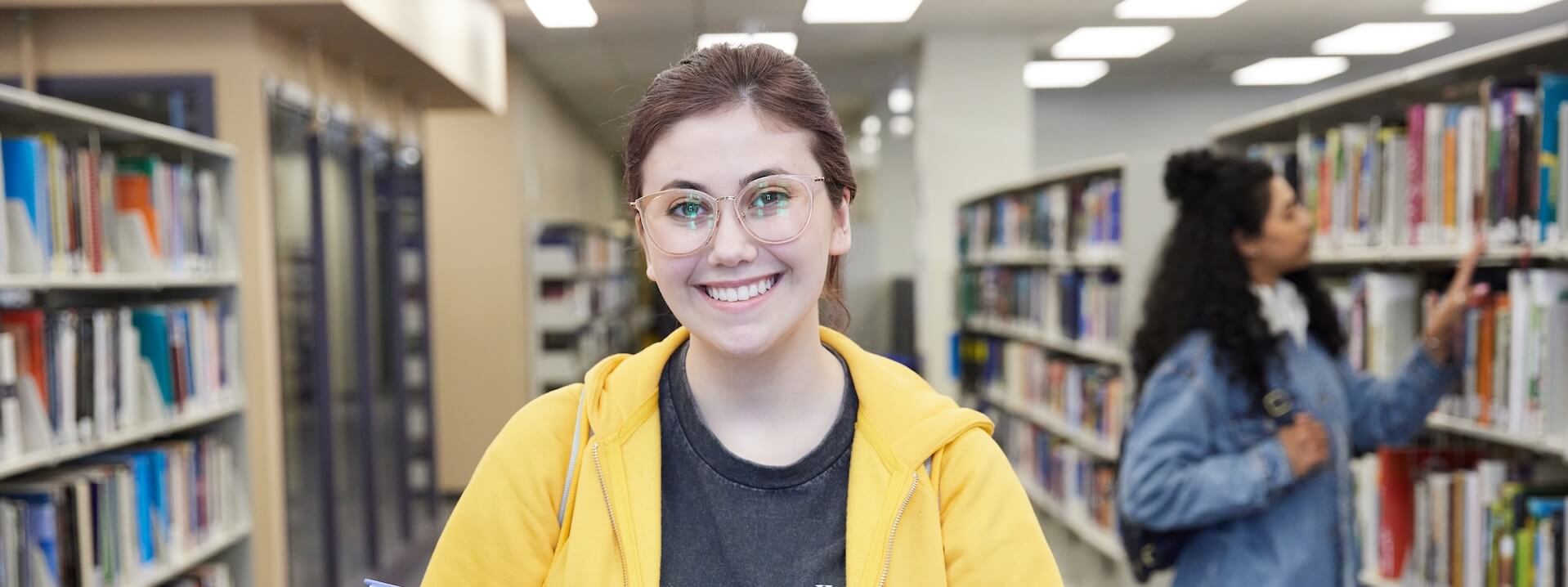Two young women collaborating at a computer in a library setting. One woman is typing on the keyboard while the other smiles and watches.