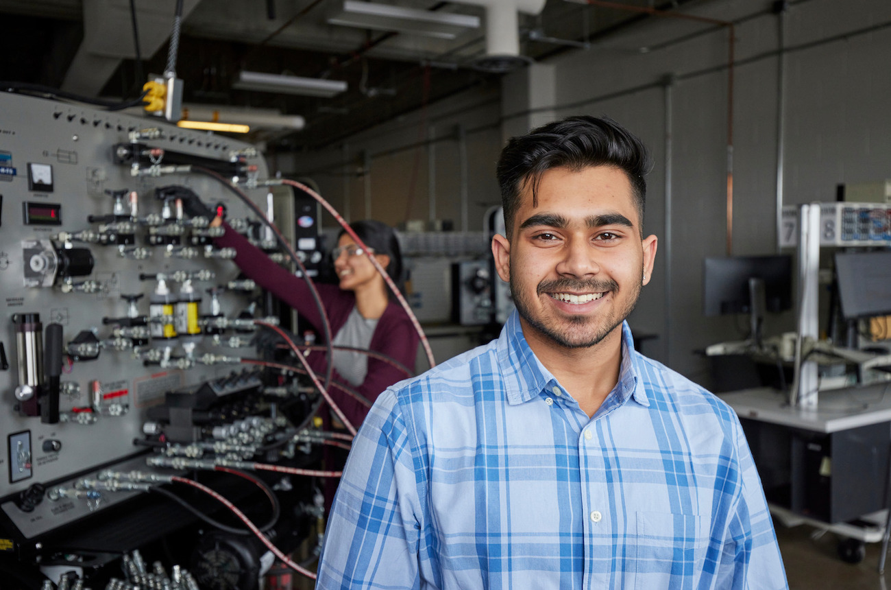 Person standing by a large window holding a phone, with a modern campus-style building visible outside.