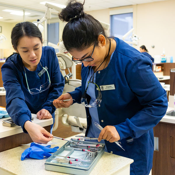 Two dental students in scrubs examining dental instruments in a clinical setting.