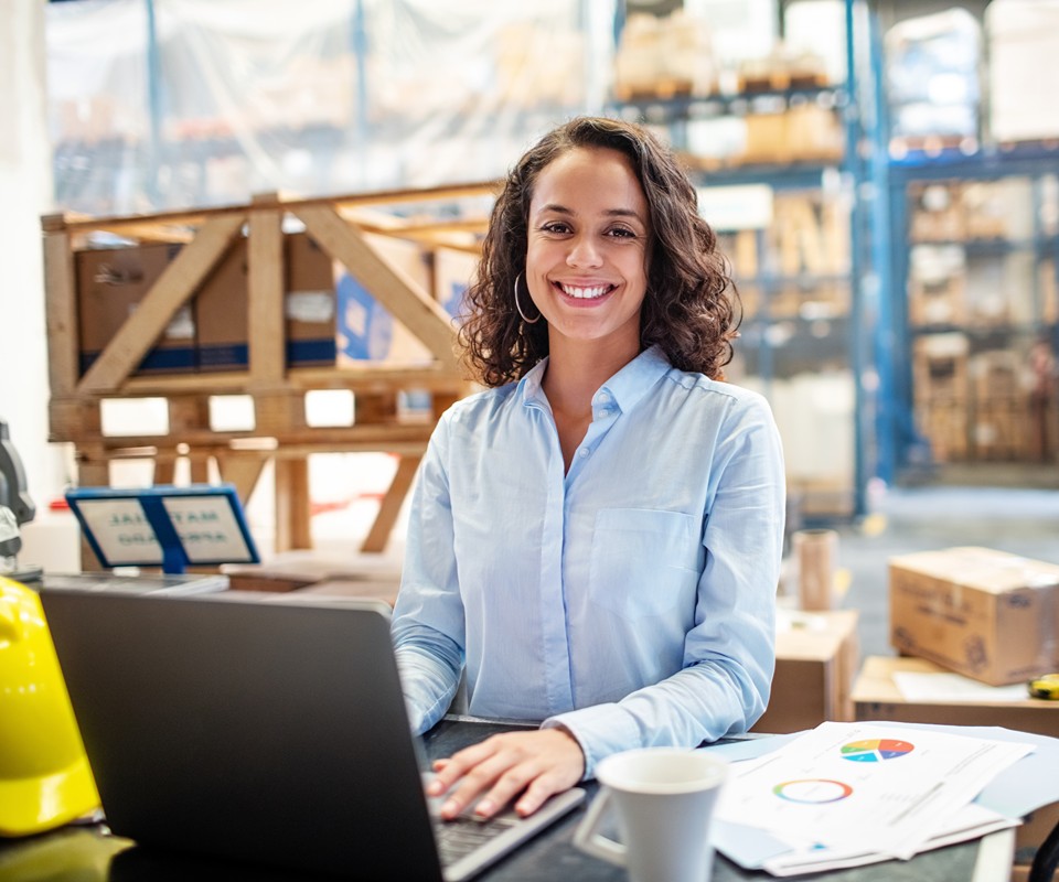 A smiling woman at a desk with a laptop, surrounded by boxes and warehouse shelving, suggesting a busy work environment.